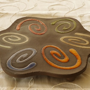 Puede incluir: Un posavasos de cerámica marrón con un diseño de espiral en tonos naranja, verde, azul y beige.