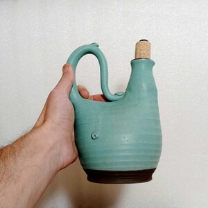 May include: A teal blue ceramic bottle with a cork stopper. The bottle has a unique shape with a curved spout and a handle.