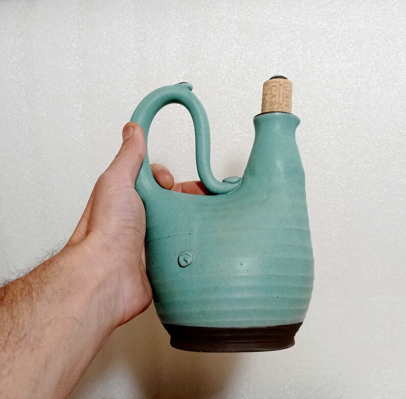 May include: A teal blue ceramic bottle with a cork stopper. The bottle has a unique shape with a curved spout and a handle.