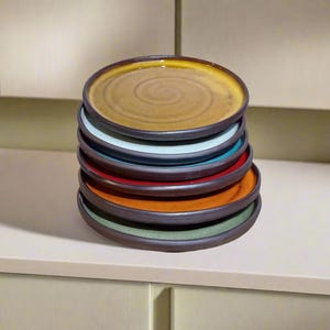 May include: A stack of eight ceramic plates in various colors, including yellow, blue, red, orange, and green. The plates are stacked on a white countertop.