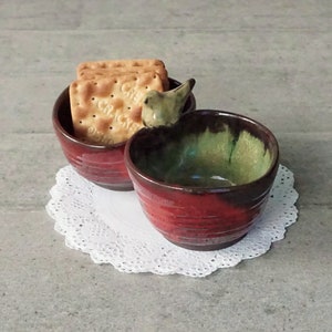 May include: Two red ceramic bowls with a green glaze on the inside. The bowls are stacked on top of each other and have a small bird figurine on the top. The bowls are sitting on a white doily.