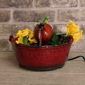 Könnte beinhalten: Eine rote Keramikfontäne mit einem Granatapfel-Design und einem Wasserfall. Die Fontäne ist von gelben Blumen und grünen Blättern umgeben.