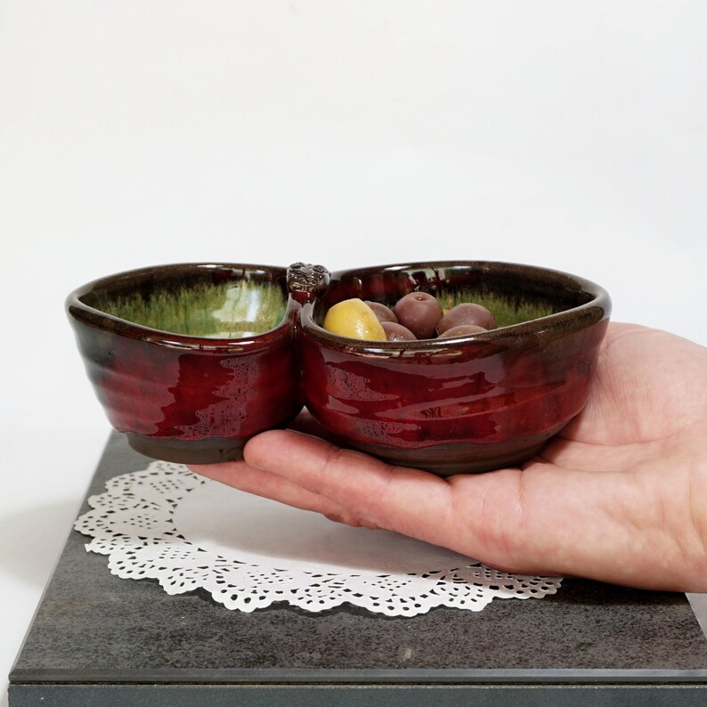 May include: A red and green ceramic divided serving dish with two sections. The dish is filled with olives and is being held in a hand.