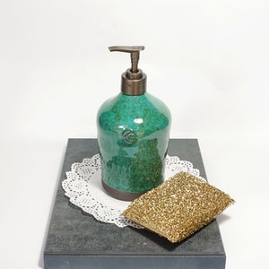 May include: A teal green ceramic soap dispenser with a brass pump top. The dispenser is sitting on a black slate surface with a white doily and a gold sponge.
