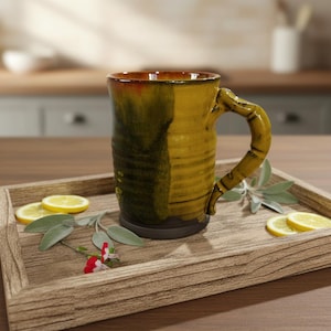 May include: A handmade ceramic mug with a distinctive, textured pattern. The mug has a mix of yellow and green tones, with a brown interior and handle. It rests on a wooden tray with lemon slices and foliage.