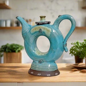 May include: A teal blue ceramic teapot with a unique donut-shaped design and a brown base. The teapot has a lid with a white floral pattern.