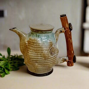 May include: A ceramic teapot with a light brown and green glaze. The teapot has a unique, textured surface and a wooden handle.