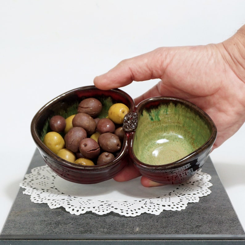 May include: A two-section ceramic serving dish with a heart shape. The dish is glazed in shades of brown, green, and black. The dish is filled with green and brown olives.