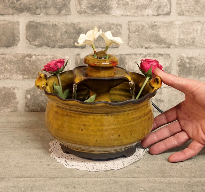 May include: A ceramic tabletop fountain with a brown and yellow glaze. The fountain has three tiers and features a cascading waterfall. The fountain is decorated with white and pink flowers.