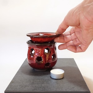 Pode incluir: Queimador de óleo de cerâmica vermelha com um design decorativo e um prato superior removível. Uma vela de chá está colocada em uma superfície preta ao lado do queimador.