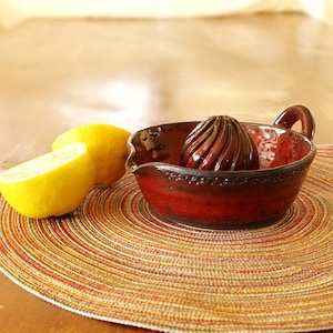May include: A red ceramic citrus juicer with a ribbed reamer and a handle. The juicer is sitting on a woven placemat with two lemons beside it.