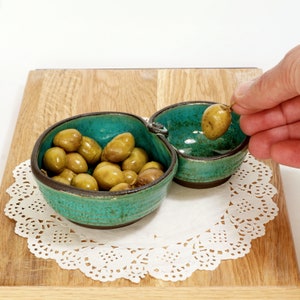 May include: A teal ceramic serving dish with two sections. One section is filled with green olives. The dish is on a white doily.