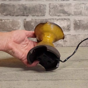 May include: A yellow ceramic vase with a black base and a cord attached to the bottom. The vase is being held by a hand.