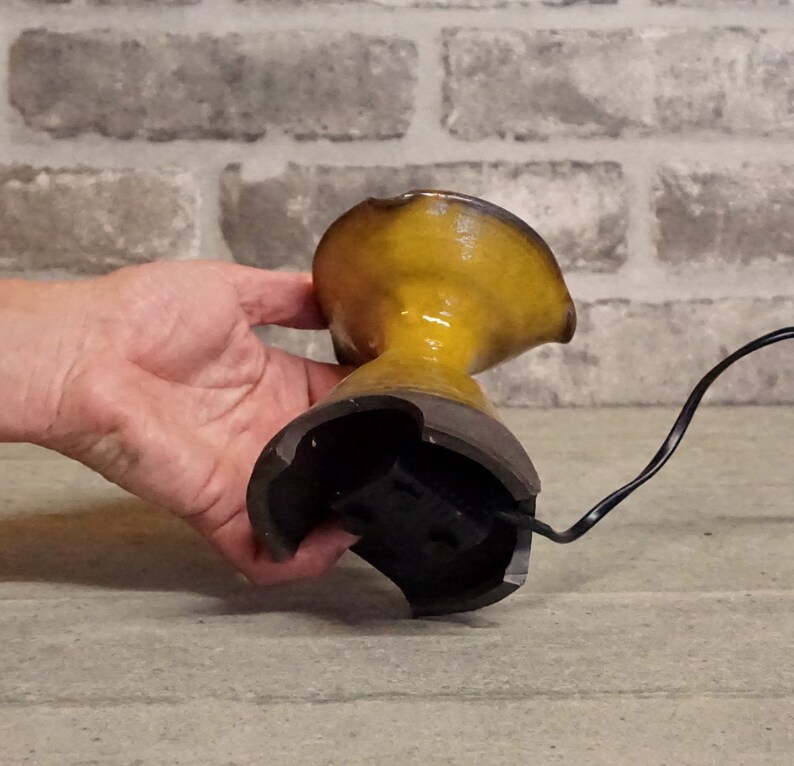 May include: A yellow ceramic vase with a black base and a cord attached to the bottom. The vase is being held by a hand.