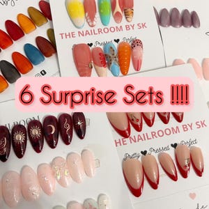 May include: Multiple sets of almond-shaped press-on nails displayed on white cards. Designs include solid colours, French tips with red cherries, celestial motifs with suns and moons, and textured 3D accents. Text on cards reads THE NAILROOM BY SK and 6 Surprise Sets !!!!