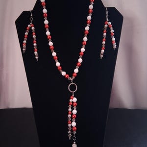 May include: A red and white beaded necklace and earring set. The necklace features a long strand of alternating red and white beads, with a silver pendant and dangling beads. The earrings have a similar design, with red, white, and silver beads.