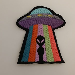 May include: Embroidered patch featuring a black alien with glowing red eyes standing in front of a rainbow beam emanating from a purple and grey UFO.