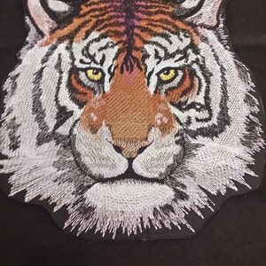 May include: Embroidered tiger face patch with white, orange, and black fur. The tiger has yellow eyes and a white muzzle.
