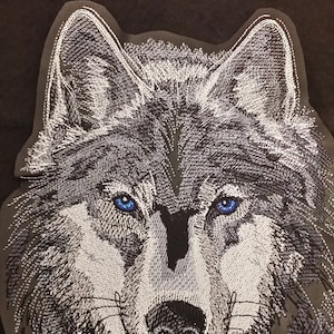 May include: Embroidered wolf head patch with a white and grey fur design and blue eyes. The wolf is looking directly at the viewer.