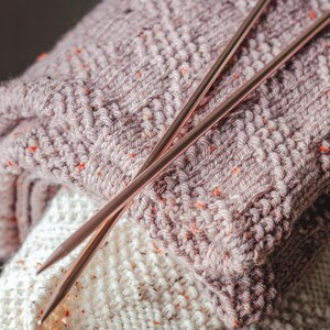 Puede incluir: Primer plano de una manta de punto en tonos malva y crema, salpicada de motas naranjas. Dos agujas de tejer de color cobre descansan sobre la manta, lo que sugiere un proyecto artesanal. La tela texturizada y los tonos cálidos evocan una sensación acogedora y artesanal.