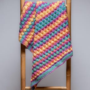 Könnte beinhalten: Eine handgestrickte Decke mit einem strukturierten, blasenartigen Muster. Die Decke zeigt eine lebendige Mischung aus rosa, gelben, blauen und grünen Farbtönen. Sie ist über eine Holzleiter drapiert.