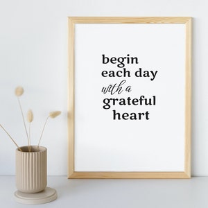 Puede incluir: Una impresión enmarcada con el texto "begin each day with a grateful heart" en letras negras sobre fondo blanco. El marco es de madera clara.