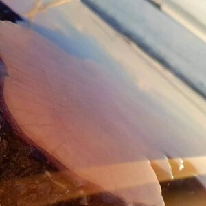 May include: A close-up of a wooden table top with a clear epoxy resin finish. The resin has a light blue and pink hue, creating a unique and beautiful effect. The wood is a dark brown color and has a natural grain pattern.
