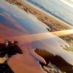 May include: A close-up of a wooden table top with a clear epoxy resin finish. The resin has a rippled effect, creating a unique and natural look. The table is set against a sandy beach background with the ocean in the distance.