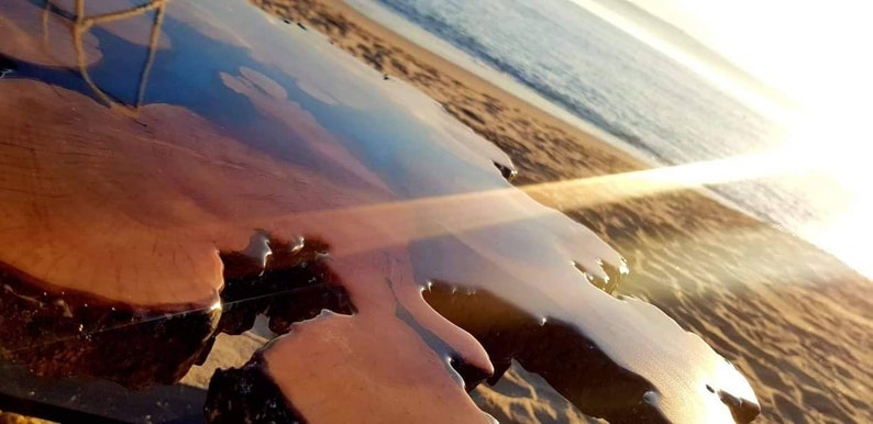 May include: A close-up of a wooden table top with a clear epoxy resin finish. The resin has a rippled effect, creating a unique and natural look. The table is set against a sandy beach background with the ocean in the distance.