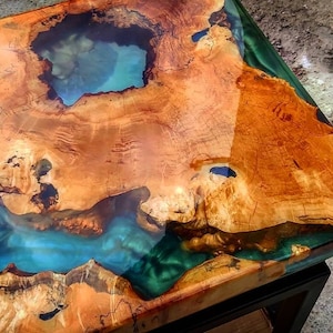 Puede incluir: Una tapa de mesa cuadrada de madera con una gran incrustación de resina azul verdosa irregular. La resina tiene un acabado profundo, claro y brillante. La madera es de color marrón rojizo cálido con un acabado natural y rústico.