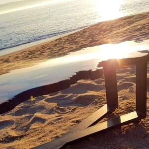 May include: A large, irregular shaped coffee table with a natural wood base and a clear epoxy resin top. The table is sitting on a sandy beach with the ocean in the background.