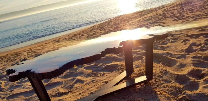 May include: A large, irregular shaped coffee table with a natural wood base and a clear epoxy resin top. The table is sitting on a sandy beach with the ocean in the background.