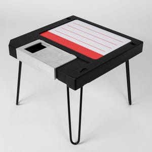 May include: A black side table shaped like a floppy disk with a red label and silver metal insert. The table has four thin black metal legs.