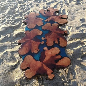 May include: A wooden table top with a blue resin inlay. The wood has a natural brown color and the resin has a swirling pattern. The table top is sitting on a sandy beach.