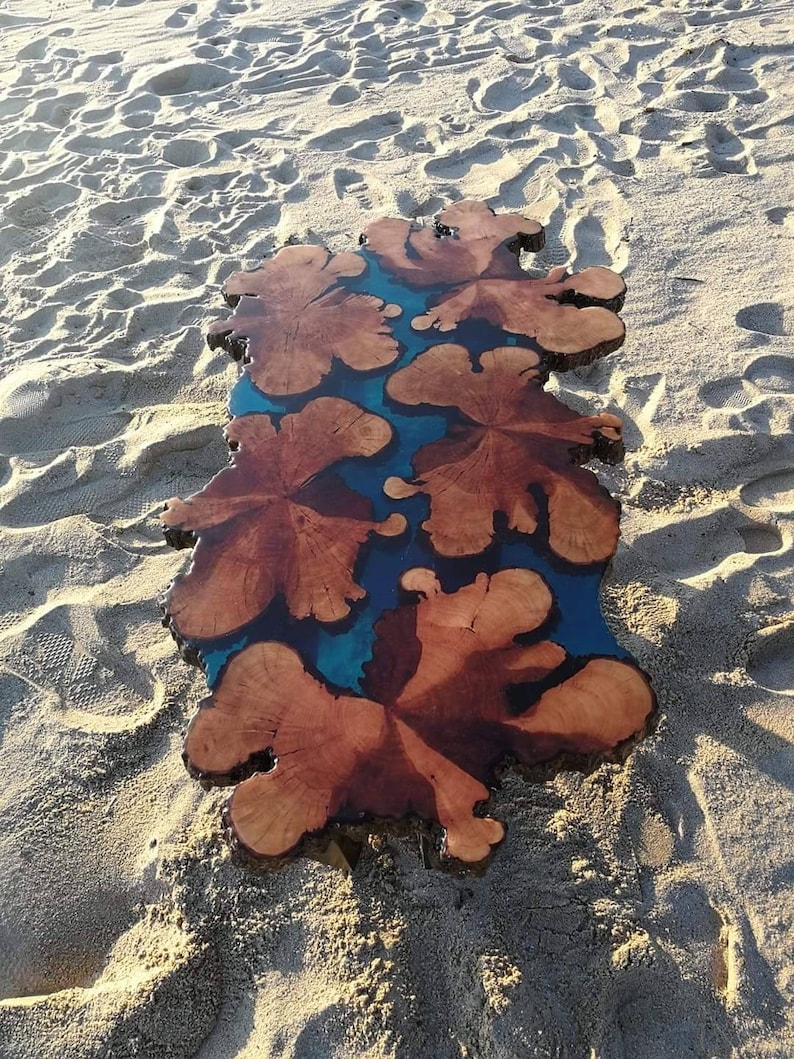 May include: A wooden table top with a blue resin inlay. The wood has a natural brown color and the resin has a swirling pattern. The table top is sitting on a sandy beach.