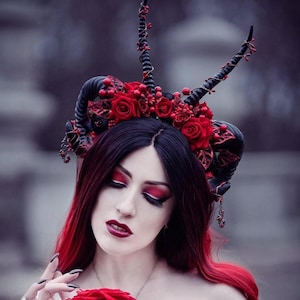 May include: A woman with black hair with red highlights, wearing a black and red dress adorned with roses. She wears a horned headpiece with red roses and berries. She holds a red rose, with dark makeup and a pendant.
