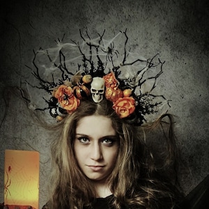 May include: A young woman dressed in a black and orange costume with a skull and flower crown. She is holding a lit candle with a spider design. The woman is posing in a dark room with a pumpkin in the background.
