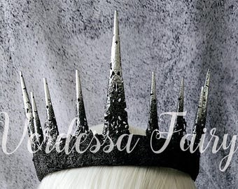 May include: A black and silver crown with tall, pointed spikes. The crown sits atop a white wig on a mannequin head. The image includes the text "Videssa Fairy" and red flowers.
