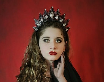 May include: A young woman with long, curly brown hair wears a black gown and a silver and black crown. She is posing with her hand on her chin, looking directly at the camera. The background is a solid red.