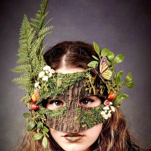 May include: A masquerade mask featuring a textured brown bark finish covered in green moss, accented with green fern fronds, small white mushrooms, orange berries, and a monarch butterfly. The mask covers the eye area with organic forest elements.