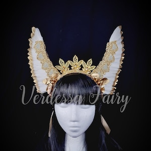 May include: A gold and white feathered headband with a gold crown and pearl accents. The headband is worn on a mannequin with black hair.