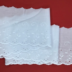 May include: White cotton lace trim with a scalloped edge and embroidered floral design. The lace is shown folded in three layers.