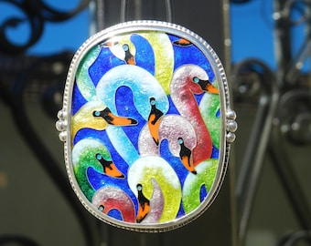 Op de afbeelding: Ovale hanger met een zilveren frame, met een kleurrijk emaille ontwerp van zwanen. De zwanen hebben witte, gele, groene en rode veren, met oranje snavels en zwarte ogen, tegen een blauwe achtergrond.