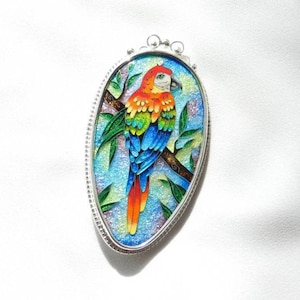 May include: Oval pendant with a colorful parrot design. The parrot features vibrant red, orange, yellow, green, and blue feathers, perched on a brown branch with green leaves. The pendant has a silver-colored frame.