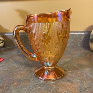 May include: An iridescent orange glass pitcher with a handle and a pedestal base. The pitcher has a floral design and vertical ribbed texture. The rim and handle have a glossy finish. The pitcher is sitting on a gray countertop.