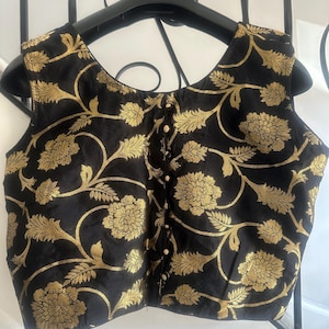 May include: A sleeveless black blouse featuring a metallic gold floral pattern throughout. The garment has a round neckline and a central vertical placket down the front, secured with a series of small, round, cream-colored buttons.
