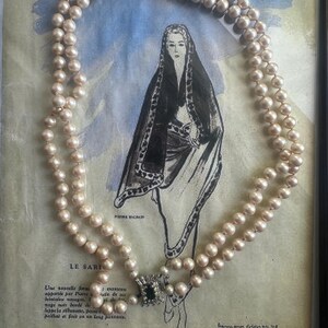 May include: A double-strand necklace of cream-colored pearls featuring a rectangular clasp with a central dark stone surrounded by clear crystals. The necklace is displayed over a vintage fashion illustration of a person wearing a dark shawl.