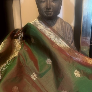 May include: A statue of Buddha draped with a green and red silk scarf. The scarf has gold floral embroidery and a gold border. The statue is made of a dark stone material.