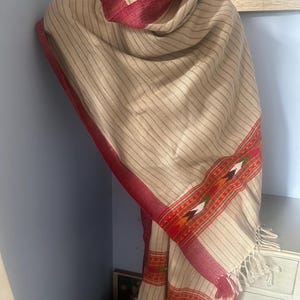 May include: Beige and red striped shawl with a decorative border. The shawl has a fringed edge and is draped over a surface. The border features geometric patterns in orange, white, and green.