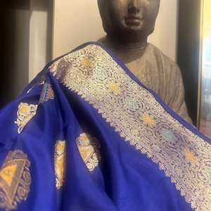 May include: A vibrant blue textile with intricate gold and teal floral patterns along the border and scattered across the fabric. The textile is draped over a statue of Buddha, creating a contrast of textures and colors.